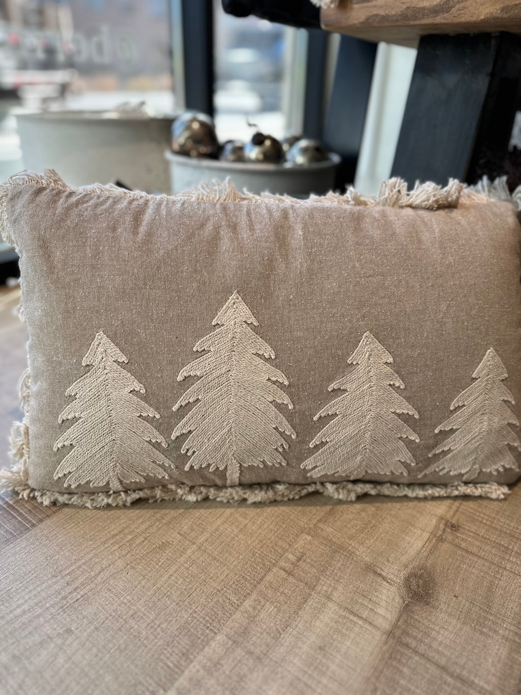 A cozy beige Christmas tree pillow with cream-colored Christmas trees on the front. The pillow is also adorned with lines of cream-colored fringe.