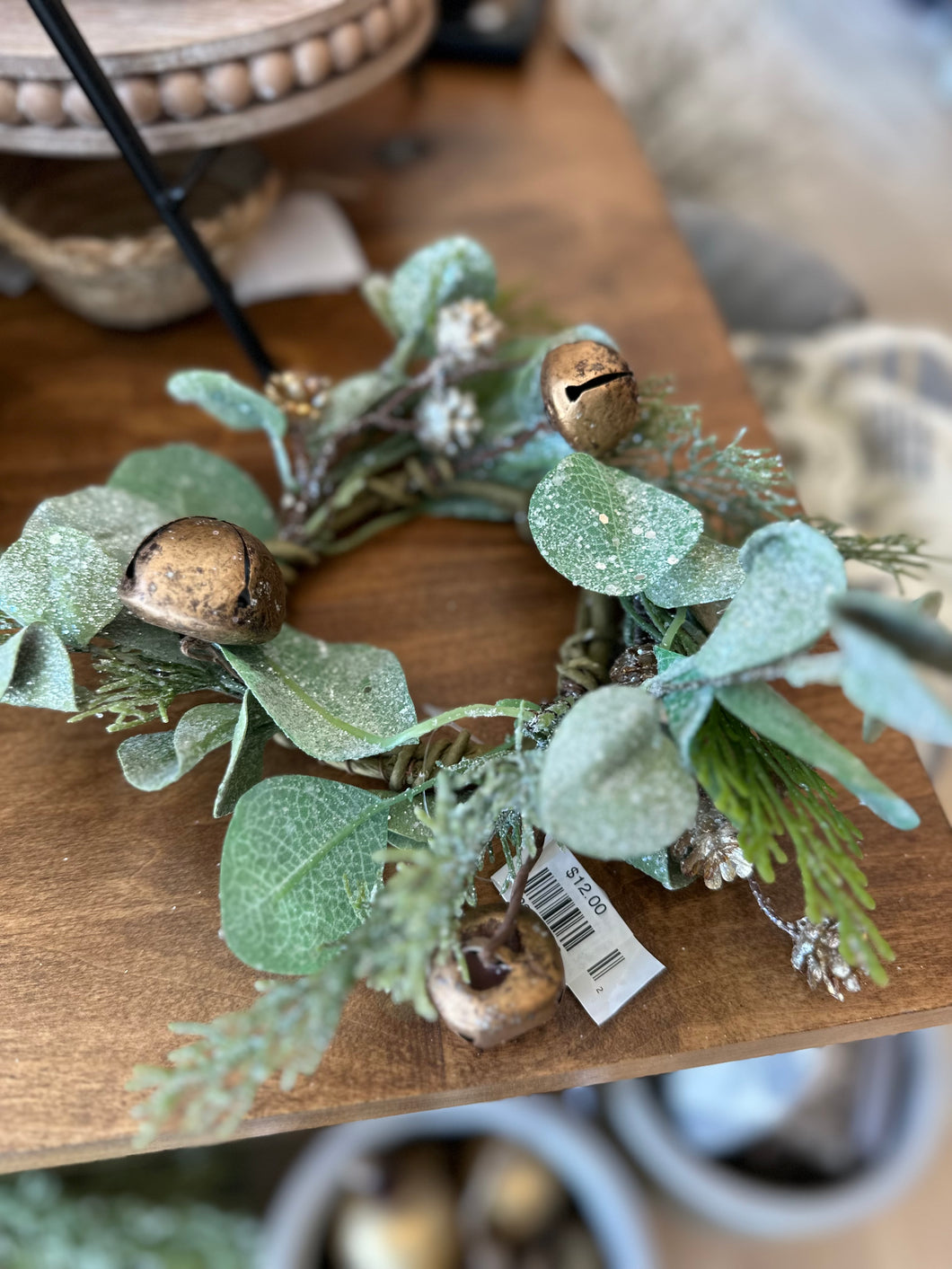 A decorative candle ring made with gold accents, eucalyptus leaves, cedar sprigs, small pine cones, and gold bells. The ring has a distressed finish and measures 10 inches in diameter with a 3-inch inner circle. It is perfect for a pillar candle.