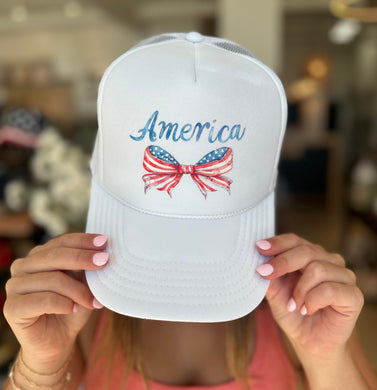 White foam trucker hat with America in blue script and a red, white, and blue coquette bow. 